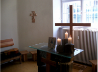 Zu sehen ist ein Altar mit drei brennenden Kerzen auf Steinen, im Hintergrund steht ein großes hölzernes Kreuz vor einem Fenster.