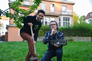 Karl und Clemens Zu sehen sind zwei Menschen im Garten, einer steht auf einem Spaten, der andere hält eine Kasse in der Hand und deutet mit dem Zeigefinger vor den Lippen an leise zu sein.